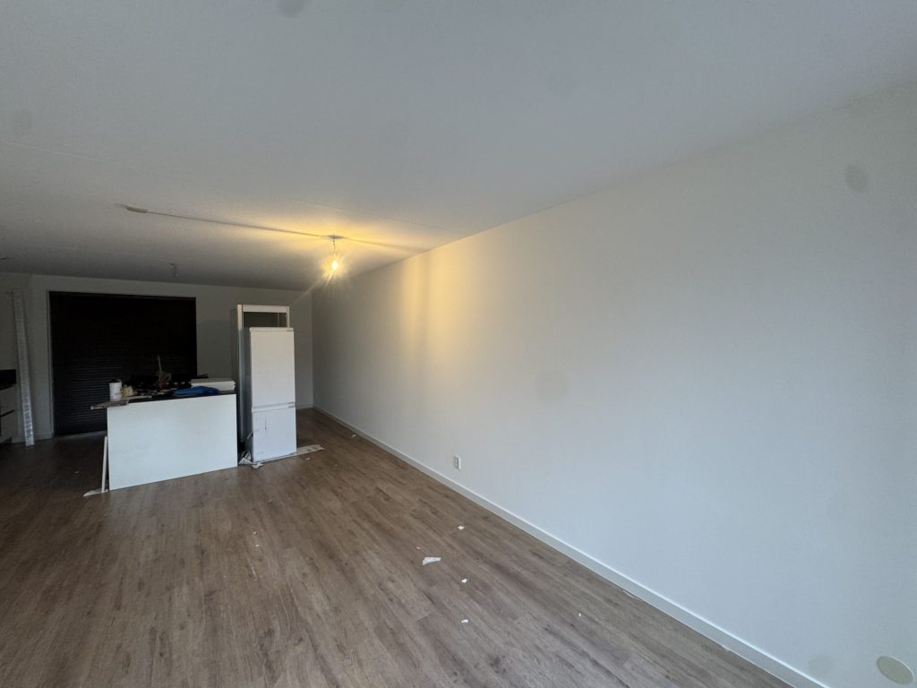 Lange, egaal wit gesausde wand in een leeg nieuwbouwappartement met houten vloer en een open keuken achterin
