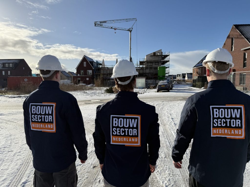Drie vakmensen van Bouwsector Nederland met witte helmen en donkerblauwe jassen lopen door een nieuwbouwwijk met een bouwkraan in de sneeuw
